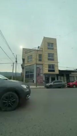 The video shows a view from inside a vehicle driving on a street in front of a three-story building. The building has a modern design with alternating horizontal bands of light-colored and darker wood or similar material. The ground floor has large glass windows with a pink advertisement inside. A gray car is parked on the side of the road in front of the building, and another dark-colored car drives past. Power lines are visible overhead. A garbage truck drives past in front of the building. There are other cars parked along the street. The sky is overcast.