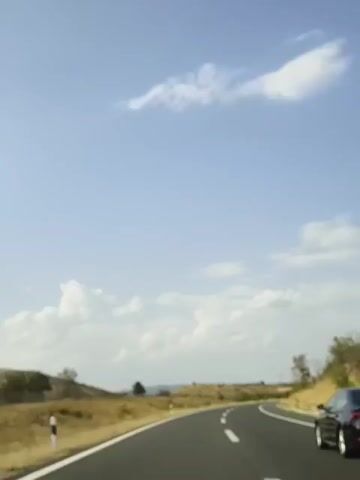 The video shows a view from a moving car on a paved road. The sky is blue with scattered white clouds. The road curves to the right. On the left side of the road, there are dry grassy hills with scattered trees. On the right side of the road, there are also hills with trees and bushes. A white line is painted along the edge of the road, and broken white lines mark the lanes. Part of a black car is visible on the right side of the frame. The time stamp indicates the video starts at 00:00 and ends at 00:04. The audio consists of a voice speaking German, with the words "Buckel" and "Schulter" audible.