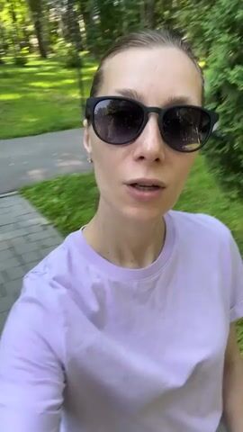 A woman wearing sunglasses and a lavender t-shirt records herself walking outdoors. She is in a park-like setting with trees and grass visible. She pauses briefly to smile at the camera before continuing her walk.