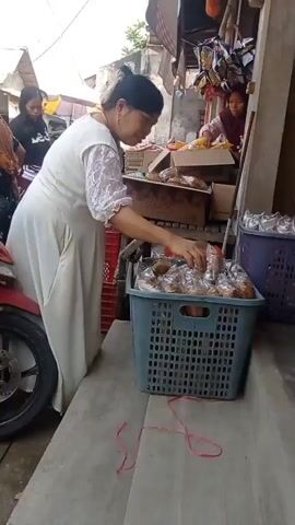 The video shows a woman in a long white dress and a black bun, reaching into a grey plastic basket filled with bagged items, seemingly at an outdoor market stall. In the background, another woman wearing a brown shirt is seated on a red motorcycle, looking towards the woman sorting the items. Further behind, other people are visible, and there are various stalls and merchandise displayed. The environment appears to be a busy marketplace with makeshift structures. The woman in the white dress is focused on her task, sorting through the goods in the basket. The red string across the bottom of the frame is a visual distraction.