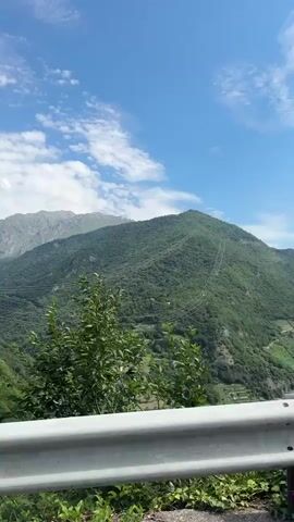 The video captures a scenic view of a mountainous landscape in Malegno, Italy. The scene is dominated by lush green mountains covered in trees, with a clear blue sky and scattered white clouds above. In the foreground, a white metal guardrail runs along the side of what appears to be a road, with some greenery and vines growing along its base.

As the camera pans, we see the vast expanse of the mountainside, dotted with small houses and buildings that form a village nestled in the valley. Power lines and a tall pylon are visible, indicating the presence of infrastructure in the otherwise natural setting. The sunlight suggests it is daytime, likely midday or afternoon, given the brightness and the shadows cast by the mountains. The overall atmosphere is serene and picturesque, showcasing the beauty of the Italian Alps. There is no visible text within the video.