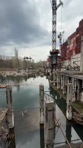 The video shows a still shot of the Chernobyl Nuclear Power Plant under a cloudy sky. The foreground features a body of water with numerous concrete pillars emerging from it. These pillars are connected by a damaged, elevated walkway. Reflections of the structures above can be seen in the water. In the midground stands a tall crane with a cab and extending arm, positioned in front of a large, dilapidated building. This building has a reddish-brown exterior and appears to be structurally unsound, with exposed concrete columns and beams. The background includes sparse trees and other industrial structures, all under the overcast sky.