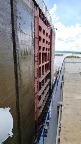 The video shows a large ship passing through a lock gate. The gate is red and appears to be made of metal. The ship is a dark color and is partially submerged in water. The water is dark and choppy. The sky is cloudy and the sun is not visible. The lock gate is in the process of opening, revealing a body of water beyond. The ship is slowly moving forward into the lock chamber. The walls of the lock chamber are made of gray concrete. There is a walkway on the right side of the lock chamber, and a railing can be seen along the edge.