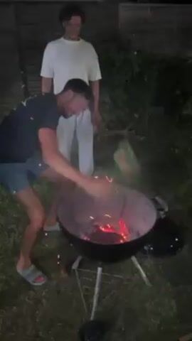 A man wearing a dark t-shirt and light shorts is seen throwing a green object into a charcoal grill that is emitting sparks. Another man stands nearby in a white t-shirt and white pants, smiling as he watches. The grill is on a grassy lawn in an outdoor setting at night, with a wooden fence visible in the background. The man adding items to the grill appears to be stoking the fire, while the other man observes with amusement.