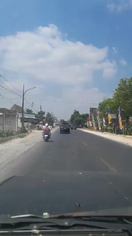 A vehicle's perspective shows a two-lane road on a sunny day. A person on a scooter rides in the left lane ahead. Two cars are visible in the distance. The right side of the road features a sidewalk, buildings, trees, and flags. The sky is blue with scattered clouds.