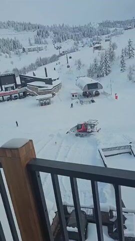 A high-angle shot from a balcony overlooking a snow-covered landscape reveals a red snowcat moving across the terrain. The snowcat is actively grooming the snow, leaving tracks behind. A small tan-colored dog is running in the snow near the snowcat. In the background, there are buildings and ski lifts covered in snow, suggesting a ski resort environment. A skier is visible near the ski lift. The balcony's dark gray railing is partially visible in the foreground.
