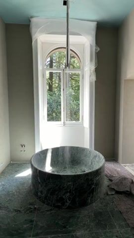 The video shows a bathroom under construction. A large, round, dark green marble tub sits in the center of the room on green marble tile flooring. A tall, arched window behind the tub is covered in plastic. The walls are a muted green color, and the ceiling is a light blue. A silver pipe hangs from the ceiling above the tub. A gray cloth lies on the floor to the right of the tub.  Water appears to be running into the tub. The window offers a view of green trees and foliage outside.