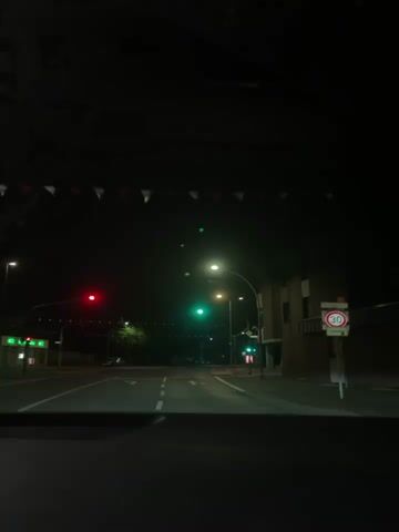 The video captures a night-time view from inside a vehicle approaching an intersection. The scene is dark, illuminated by streetlights and traffic signals.

In the foreground, the road is visible with white lane markings indicating straight-ahead and turn arrows. To the left, a traffic light is red, while a traffic light further ahead and slightly to the right is green. As the video progresses, the red light on the left also turns green. Multiple streetlights are active, casting light on the road and surroundings.

On the right side of the road, a brick-colored building is visible, and several road signs are present. A circular speed limit sign with a red border and a white background clearly shows "30" in black, indicating a 30 km/h speed limit. Another triangular warning sign is also visible on the right. Across the street, strung between poles, are decorative banners or flags, appearing as a string of small, triangular shapes.

No people are explicitly visible, and the weather appears clear. The activity is driving at night, navigating an intersection as the traffic lights change to green, permitting movement. The overall setting appears to be an urban or suburban area.