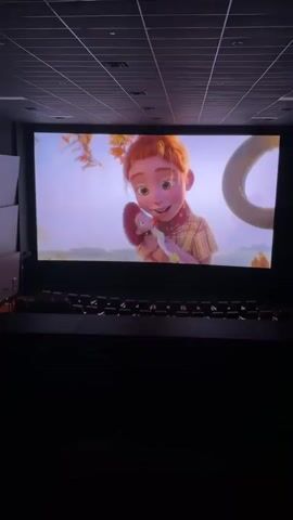 The video captures a cinema screen displaying a scene from an animated movie. In the foreground of the screen, a young girl with red hair and a plaid shirt is interacting with a stuffed toy. She is holding it up and appears to be speaking to it. The background of the scene shows a tree with autumnal foliage, a tire swing hanging from a branch, and a soft, blurred landscape. As the scene progresses, the girl is seen kneeling near the tree, then she appears to be startled and jumps away from the tire swing. The camera is positioned in the dark cinema, showing rows of empty seats in front of the screen. The lighting is dim, focusing attention on the illuminated movie screen.