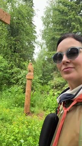The video shows a woman in sunglasses and a jacket standing on a wooden walkway in a park. She smiles and gives a thumbs-up to the camera. Behind her, a wooden sign with Cyrillic lettering hangs from a post. The walkway leads into a wooded area with lush green foliage. Another person in a red raincoat walks away from the camera on the walkway. A carved wooden post with decorative elements stands to the left of the walkway. The sky appears overcast.