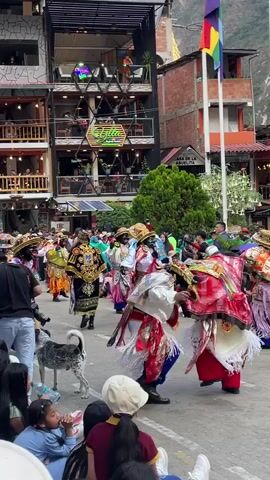 The video shows a street festival or parade in a town. In the foreground, a few people are seated on the ground, watching the event. A spotted dog is also present, walking among the seated spectators. The main focus of the video is a group of dancers in elaborate, colorful costumes. They are wearing masks that cover their faces and have large, decorative hats. Their costumes feature bright fabrics, intricate embroidery, and fringes. The dancers are moving in a coordinated fashion, likely performing a traditional dance. Behind the dancers, there are multi-story buildings with balconies and shops. A rainbow flag is visible on a flagpole. The environment appears to be outdoors, with people gathered to observe the parade. The overall scene suggests a lively cultural celebration.