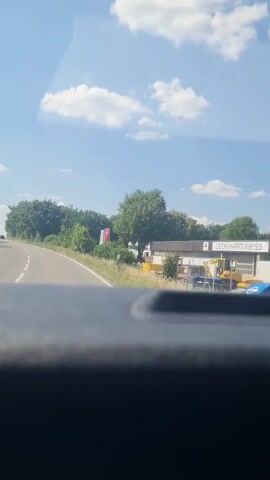 The video captures a view from inside a vehicle as it travels along a country road on a sunny day. The scene is rural, with lush green trees and vegetation lining the road. In the distance, a few buildings, including what appears to be a house with a white facade and a red roof, are visible. The sky is blue with scattered white clouds, indicating fair weather.

On the right side of the road, there are some structures that look like a business or industrial area. One building has a yellow awning, and there are construction materials and equipment, including a yellow excavator, visible. Text on a sign is partially visible, reading "LEONHART". Another prominent feature is a large red and white flag or banner displayed near the road.

The road itself is paved and appears to be a two-lane highway with a white line marking the lanes. The perspective suggests the viewer is in the passenger seat or the driver's seat of a car, looking forward through the windshield. The time of day appears to be daytime, likely late morning or early afternoon, given the bright sunlight. The overall mood is peaceful and observational, focusing on the passing scenery. There are no people clearly visible in the frame, and the activity is simply the act of driving through the landscape.