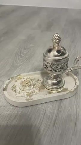 The video shows a silver incense burner on a white oval tray with gold accents, sitting on a light grey wood-patterned surface. Smoke is emanating from the burner. The tray has a decorative flower-like design on one side. The burner has a handle and a lidded top. The camera remains still.