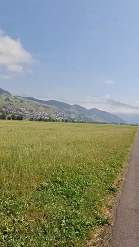 This video shows a POV shot of a person walking down a paved path through a large, grassy field. The path stretches into the distance, lined by the field on both sides. In the background, there are rolling hills and mountains, with a town or village nestled on the side of one of the hills. The sky is clear and blue with a few wispy clouds. The sun is shining, creating a bright and airy atmosphere. There are no other people or animals visible in the video. The terrain appears to be flat and open, suggesting a rural or semi-rural setting. The vegetation in the field consists primarily of tall grass and some wildflowers. The path appears to be well-maintained, though there are some minor cracks and imperfections visible. The overall impression is one of peace and tranquility, with a sense of journey and forward movement.