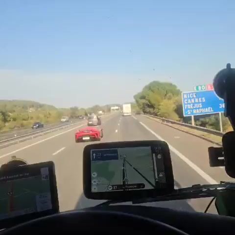 A red sports car is driving on a highway. The car has a large spoiler on the back. There are other vehicles on the highway, including a gray van and a white truck. The sky is blue and the sun is shining. The highway is surrounded by trees.