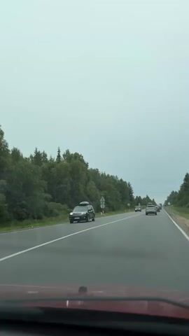 The video shows a view from inside a car traveling on a two-lane highway. The sky is overcast and gray. On the left side of the road, there is a dense forest with tall trees. On the right side of the road, there are also trees, but they appear less dense. There are several cars and a bus on the road, all traveling in the same direction as the camera car. The road surface is asphalt and appears to be in good condition. There are white lane markings on the road. There are also a few road signs visible along the side of the road. The camera car appears to be red, based on the reflection in the lower part of the frame.