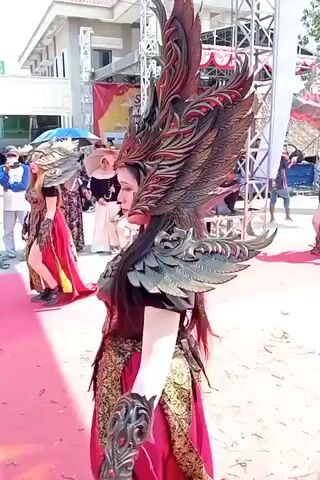 The video shows two individuals dressed in elaborate, ornate costumes that resemble mythical creatures. Both wear large, elaborate headdresses with feather-like designs and metallic armor over their shoulders and arms. Their outfits consist of dark tops and flowing red skirts with gold trim. They are walking on a red carpet outdoors, with a crowd of spectators visible in the background. Buildings and scaffolding are also present in the environment. The individuals appear to be participating in a performance or parade.