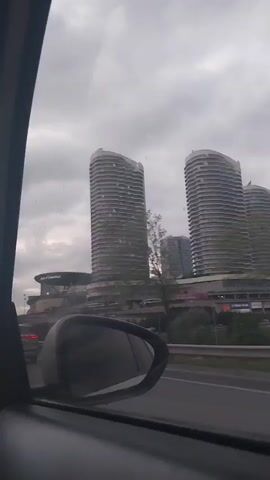 From inside a moving car, the video shows a view of the Mall of Istanbul and its adjacent high-rise buildings on a cloudy day. Other cars can be seen on the road. The rearview mirror of the car is visible in the lower left corner of the frame.
