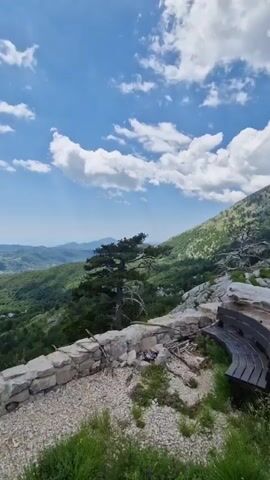 The video opens with a low-angle view of a stone wall constructed of rough-hewn rocks. Beyond the wall, a lush, green landscape unfolds, with rolling hills covered in dense trees stretching towards a hazy horizon. The sky above is a vibrant blue, dotted with fluffy white clouds that cast shadows on the terrain below. A solitary evergreen tree with a distinctive, gnarled trunk stands prominently near the center of the frame, positioned just beyond the stone wall. To the right of the wall, a dark, curved bench is visible, suggesting a viewpoint or resting spot. The ground in the foreground is a mix of gravel and patchy green grass. The camera slowly pans to the right, revealing more of the mountainous terrain and the expansive valley. The overall impression is one of a serene, natural environment, likely a scenic overlook.