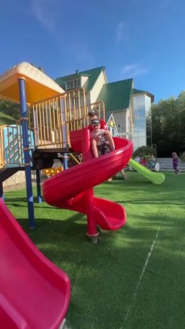 A young boy wearing a red shirt and a black baseball cap is sitting on the top of a red spiral slide at a playground. The playground has a green artificial turf surface and a bright blue sky with some clouds above. In the background, there is a multi-story building with a green roof, and other children are visible playing on a green slide further away. The boy appears to be holding a camera or phone and looking down the slide. The red slide is curved and winds downwards. Another red slide is partially visible in the foreground.