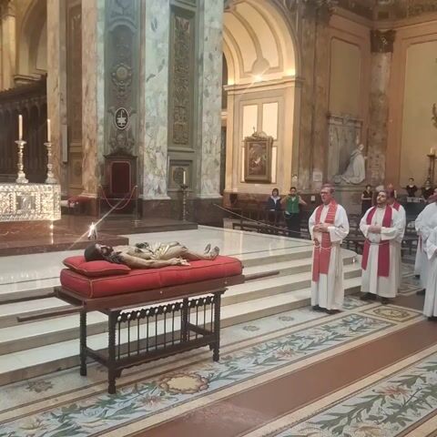 The video shows the inside of a large cathedral filled with people. In the foreground, a wooden table with a red cushion displays a recumbent statue of Jesus. Several priests in white robes with red stoles stand nearby. As the camera pans, the vast crowd of people becomes visible, many holding up their phones, seemingly to record the scene. The cathedral is decorated with marble pillars, ornate carvings, and religious paintings. The overall atmosphere suggests a religious ceremony or gathering.