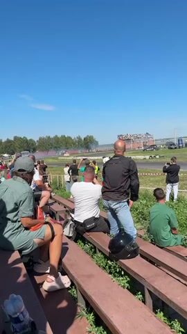 A group of spectators watch two cars drift on a sunny day from a wooden bleacher. In the foreground, several people are seated on the bleacher, some with backpacks and water bottles. A person in a green shirt is seated to the right of the frame, facing away from the camera. Behind the seated spectators, more people stand and watch the event. A black helmet sits on the bleacher in the middle of the frame. In the middle ground, two cars are drifting on a paved track. Smoke billows from the tires of the drifting cars. A yellow truck is parked in the background to the right of the track. A control tower with a white structure on top stands in the distance to the right. The sky is clear and blue.