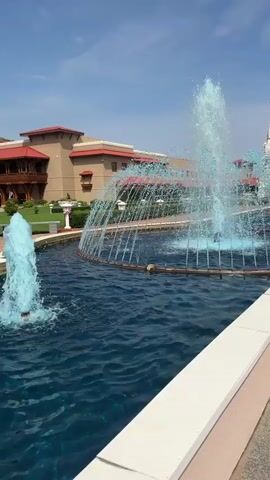 A video displays a wide shot of a fountain with multiple jets of water spraying into the air. The fountain is located in a large, blue pool of water. In the background, a white building with multiple spires and domes, resembling a temple, stands under a clear blue sky. A smaller, brown building with red trim is also visible to the left of the temple. The scene appears to be outdoors, possibly a public space or religious site.