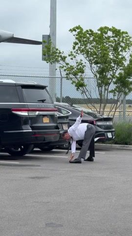 A person, dressed in a white dress shirt and grey pants, is bending over to pick up something from the ground between two parked cars. The scene is outdoors in a parking lot, with a chain-link fence and trees in the background. The sky appears overcast, suggesting it is daytime with possibly cloudy weather. One of the cars is a black SUV, and the other is a black sedan. A portion of an aircraft wing is visible in the upper left corner of the frame. The visible license plate on the SUV reads "ELITE839". The setting appears to be in the Town of Southampton, United States of America.