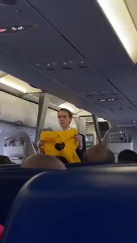 Here is a description of the video:

The video shows a male flight attendant in a suit standing in the aisle of a commercial airplane and holding up a yellow life vest. The life vest has black lettering on it. Passengers are seated on either side of the aisle. Some have their heads visible and are looking at the flight attendant. The airplane's interior is visible, including overhead compartments, reading lights, and seat backs. There are newspapers in the seatback pockets.
