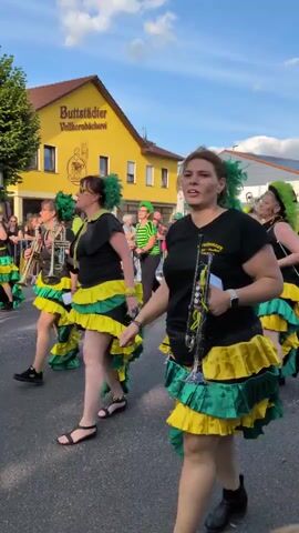The video shows a parade on a street with buildings and trees in the background. A group of women dressed in black tops and ruffled skirts in yellow and green are marching. Some are holding trumpets. A woman in the foreground on the right is holding a trumpet and looking to the left. Another woman to her left is pointing with her left hand. In the background, there are more people, some wearing green hats and vests, and a person pushing a stroller. A yellow building with a sign that says "Buttstädter Vollkornbäckerei" is visible. The sky is visible and appears to be partly cloudy.