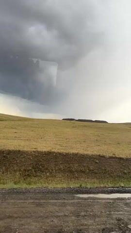 The video shows a panoramic view of a vast, open landscape under a stormy sky. The terrain is characterized by rolling hills covered in dry, golden grass. A dirt road with puddles of water runs along the bottom of the frame, suggesting recent rain. In the distance, the land rises to form a plateau or mesa with a dark, rocky top. The sky is filled with heavy, dark clouds, with patches of lighter sky and sunlight breaking through in some areas, creating a dramatic and somewhat ominous atmosphere. The camera pans slowly across the scene from left to right. There are no people or animals visible in the video. The overall impression is one of a remote, natural environment experiencing unsettled weather.