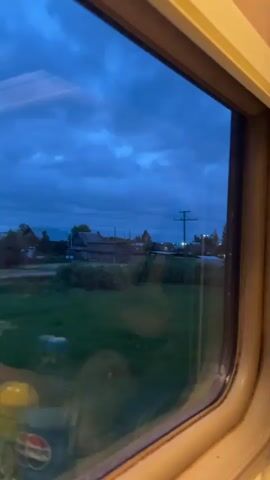 A close-up shot through a train window, tinted with reflections, reveals a rural scene at dusk. The overcast, blue sky looms above a landscape marked by scattered wooden houses and lush green fields. Utility poles punctuate the view, carrying power lines across the horizon. As the train moves, cars can be seen driving through a village road. The interior reflections on the glass obscure the full clarity of the external view.