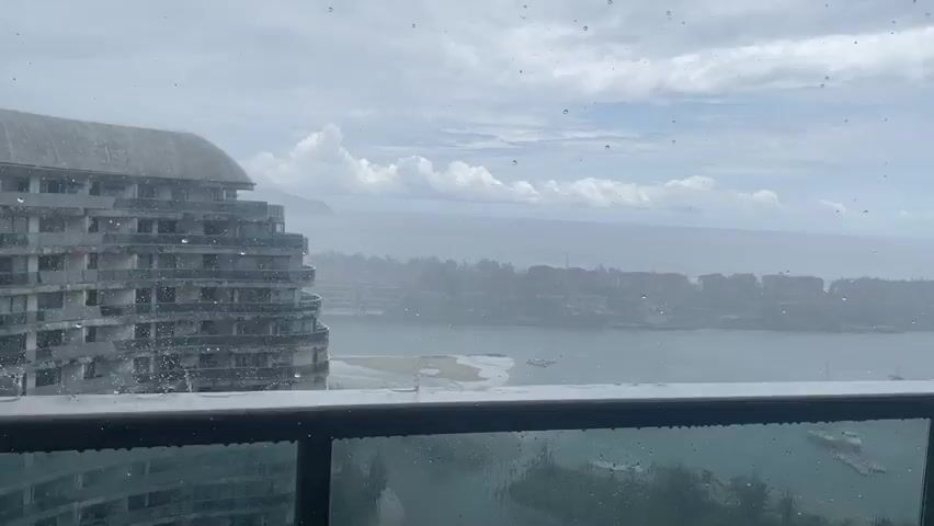 A drone shot looks out from a balcony with a glass railing onto a cloudy day with rain streaking down the glass. In the foreground, the railing is visible, and beyond it, an unfinished, multi-story building with a curved facade is prominent on the left. In the middle ground, there's a body of water, possibly a bay or ocean, with a shoreline featuring trees and buildings. A sandy area with a few boats is visible in the water. The background shows the ocean meeting a hazy horizon under a sky filled with large, grey clouds and patches of white. The overall impression is a bleak, overcast day with inclement weather.
