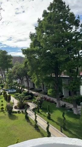 The video pans across a resort-like setting with lush green landscaping and buildings. In the foreground, a white railing is visible. The camera moves from left to right, revealing well-maintained gardens with manicured hedges, curved pathways, and tall trees. Buildings with multiple stories and balconies are interspersed among the trees. The sky is a clear blue with scattered white clouds. In the distance, the sea is visible. The overall impression is one of a tranquil and well-kept outdoor environment. No people are clearly visible, although a figure might be glimpsed on a balcony in the middle distance.