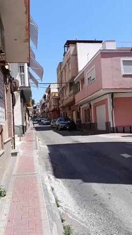 The video shows a view down a narrow street in what appears to be a residential area. The street is paved and lined with buildings of various heights and colors, including white, beige, and pink. Cars are parked along the street on both sides. The sidewalk on the left is paved with red and gray bricks. The sky is clear and blue, suggesting a sunny day. No people are clearly visible in the main view of the street, but there appears to be a figure walking towards the end of the street. The video is taken from a stationary position on the sidewalk.