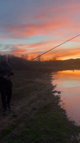 A man is fishing at a body of water at sunset. The sky is a mix of pink, orange, and blue. The water reflects the colors of the sky, and there is a hill in the background. The man is standing on the shore, and he is holding a fishing rod. A vehicle appears to be parked near the water on the other side.