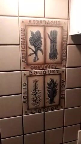 The camera pans over a tiled wall, showing two groupings of decorative tiles. The tiles are arranged in a two-by-two square. The top set of tiles depicts asparagus and artichokes. The bottom set of tiles depicts garlic and lavender. The tiles are arranged in a pattern with the tile subject in the center and the words related to the image arranged around the borders. The wall is mostly beige with white tiles.