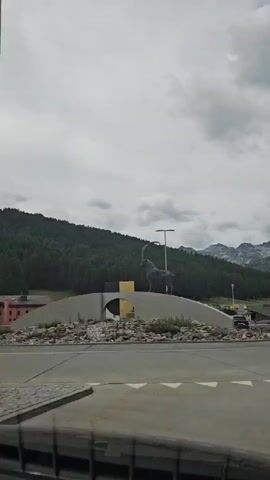 A statue of an ibex with large, curved horns stands atop a concrete structure resembling a bridge or ramp. The structure is decorated with rocks and debris. Behind the statue, a dense green forest covers a mountain slope. The sky is cloudy. The footage appears to be captured from inside a vehicle, as indicated by a reflection on the lower part of the screen.