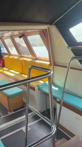 The video shows the interior of a boat with yellow and turquoise seating. A blonde woman wearing sunglasses, jeans and a black jacket is sitting on one of the seats. The boat is moving on water, and through the windows, we see a scenic view of a lake with mountains in the background and a small dock. The interior of the boat includes wooden tables, steel railings and stairs, and a black-and-grey patterned floor.