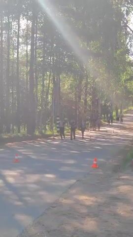 The video shows a group of people running on a paved path in a forest. The sun is shining through the trees, casting shadows on the path. There are orange cones on the side of the path, marking the course. The runners are wearing athletic clothing and some have backpacks. The environment is a forest with tall trees. The activity appears to be a race or a training run.
