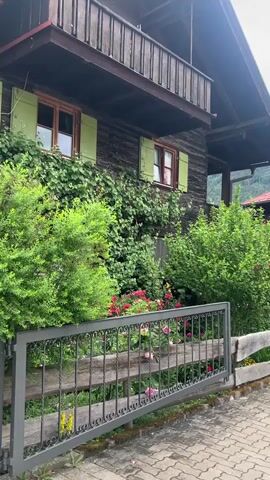 The video captures a serene street scene in what appears to be a traditional Alpine village, likely Oberstdorf, Germany, given the context.

The initial shot focuses on a dark wooden chalet-style house with green shutters on its windows and a wooden balcony on the upper floor. The facade of the house is largely covered in lush green climbing plants. In front of this building, a vibrant garden bursts with green bushes and numerous red and pink roses. A decorative dark gray wrought iron gate stands in the foreground, separating the garden from a paved path made of interlocked paver stones. A wooden fence extends from the gate.

As the camera slowly pans to the right, a second, larger building of similar traditional design is revealed. This multi-story structure also features dark wooden siding, a red-tiled roof, and multiple balconies. These balconies are distinctively decorated with bright yellow awnings and flower boxes brimming with colorful blooms. On the ground level of this building, a green sign with the word "Alban" in white letters is visible above an entrance. To the right, a small paved area includes a glimpse of a parked vehicle.

The sky is overcast and bright, indicating a daytime setting with cloudy weather. The scene is characterized by abundant greenery, traditional architecture with decorative elements, and a sense of quiet charm. No people are visible, and the only discernible activity is the camera's panning motion.