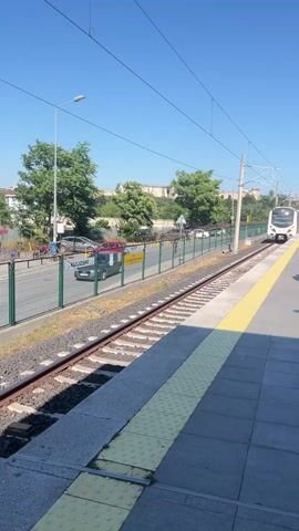 A train station platform with a train approaching on the tracks. The train is white and silver and has a green section on its side with advertisements. There are people on the platform waiting for the train. There are trees and buildings in the background. The sky is clear and blue.