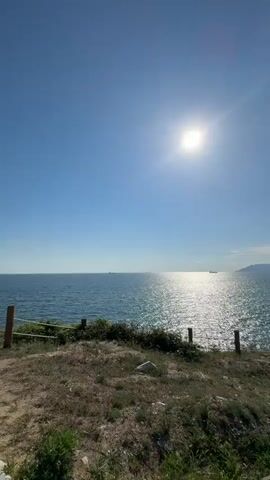 The video captures a scenic view of the sea on a bright, sunny day. The sun is high in the clear blue sky, casting a strong glare on the water. The sea is a deep blue, with gentle waves reflecting the sunlight. In the distance, several ships can be seen. The foreground shows a grassy cliff overlooking the sea, with a simple wooden fence and posts along the edge. A paved pathway made of irregular stone slabs winds through the grassy area. There are some scattered rocks and small bushes along the path and on the cliff. The overall impression is one of a peaceful and beautiful coastal landscape.
