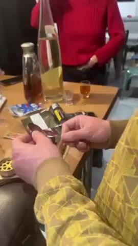The video shows a person sitting at a table and rolling what appears to be a marijuana cigarette. The person wears a tan shirt with a yellow geometric pattern. The table has several items on it, including two bottles of liquid, two small glasses of liquid, a pack of rolling papers, a partially rolled marijuana cigarette, a lighter, a cigarette, a tin, and a phone. The background includes other people sitting at a table. One person is wearing a red sweater. The environment looks like someone's dining area. The table is wooden.