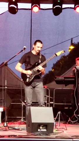 A man is playing a black electric guitar on a stage. He is wearing a black t-shirt and grey pants. There are stage lights to the left of the frame, some of which are red and white. There is a microphone stand in front of him. In the background, there is a keyboard, a speaker, and what appears to be a tent or canopy. The sky in the background is a dusky purple, suggesting it is evening.