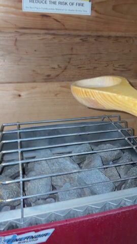 Here is a description of the video:
A wooden ladle pours water onto heated sauna rocks. The rocks sit inside a metal cage on top of a red heater. A sign above reads, "REDUCE THE RISK OF FIRE." The sauna walls are made of wood.