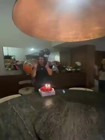 A group of people is gathered around a table, clapping and singing, presumably celebrating a birthday. A cake with a lit candle sits on the table. The room has warm lighting and appears to be a restaurant or private dining space, with wooden accents and modern decor. The people are mostly women and appear to be happy and engaged in the celebration.