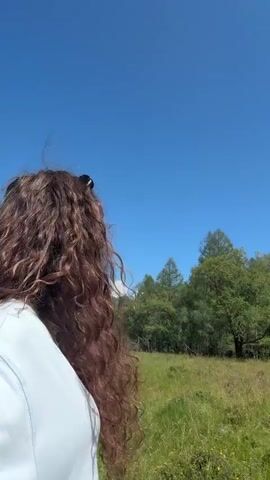 The video opens with a woman from the back, facing away from the camera, with long, wavy brown hair and sunglasses on her head. She is standing in a grassy field with trees in the background. She turns her head to look directly at the camera, revealing her face. She is wearing a light blue sweatshirt. The scene is bright and sunny with a clear blue sky. The camera then pans up to show the trees and sky, with a long, cylindrical wooden structure with multiple circular openings visible among the trees. The sun is shining brightly, casting a lens flare. The camera continues to pan across the trees and the grassy field, showing more of the forest and the clear blue sky. The video concludes with a wide shot of the trees and the sunny sky.