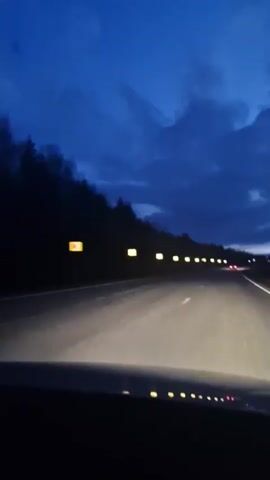 A dashcam perspective shows a car driving on a multi-lane highway at dusk or early night. The sky above is a deep blue with visible clouds, indicating twilight. The car's headlights illuminate the asphalt road ahead, highlighting white lane markings. To the left, a dark silhouette of a forest lines the road. Along the side of the road, several rectangular lights with a yellow hue are visible, likely marking road signs or structures. In the distance, the red taillights and white headlights of other vehicles can be seen. The dashboard of the car is visible at the bottom of the frame, showing various illuminated indicators. The scene evokes a sense of travel and movement in the quiet evening hours in the area of Dybki, Russia. There is no discernible text visible in the video frame.