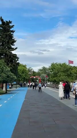 The video shows a wide pedestrian walkway bordered by a blue bike lane on the left and trees and people on the right. Several people are walking along the pathway, some in small groups and others individually. In the background, there is a red archway with some text on it. Turkish flags are visible on poles in the distance. The sky is blue with scattered clouds, and the overall setting appears to be a park or recreational area on a pleasant day.
